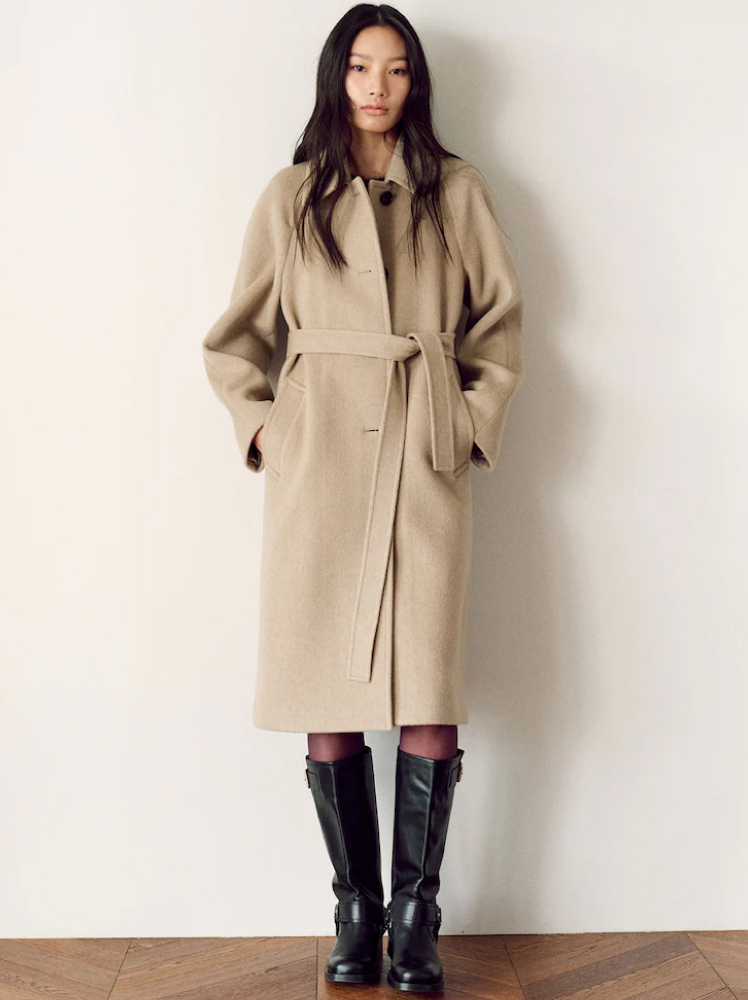 A model poses with her hands in her pocket while wearing a long beige coat that buttons up down the center and has a belt around the waist. The coat is paired with sheer black tights and a pair of knee-length leather riding boots with buckle detailing.