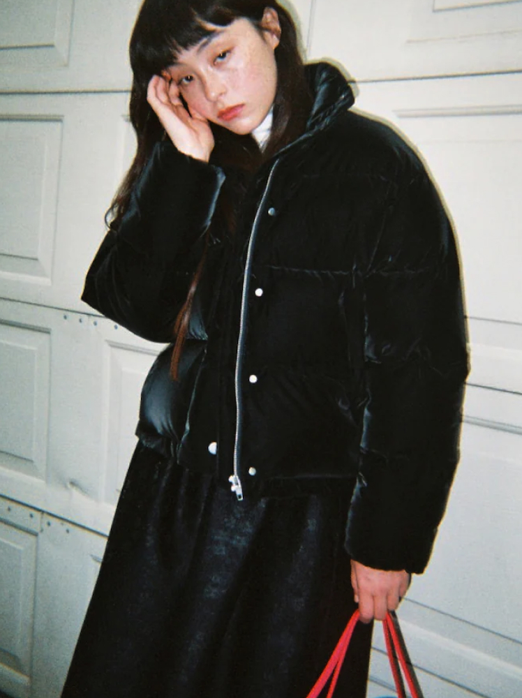 A woman leans up against a white garage door and rests her right hand on her face while wearing a velvet-like black puffer jacket and an A-line black skirt and is holding a red bag.