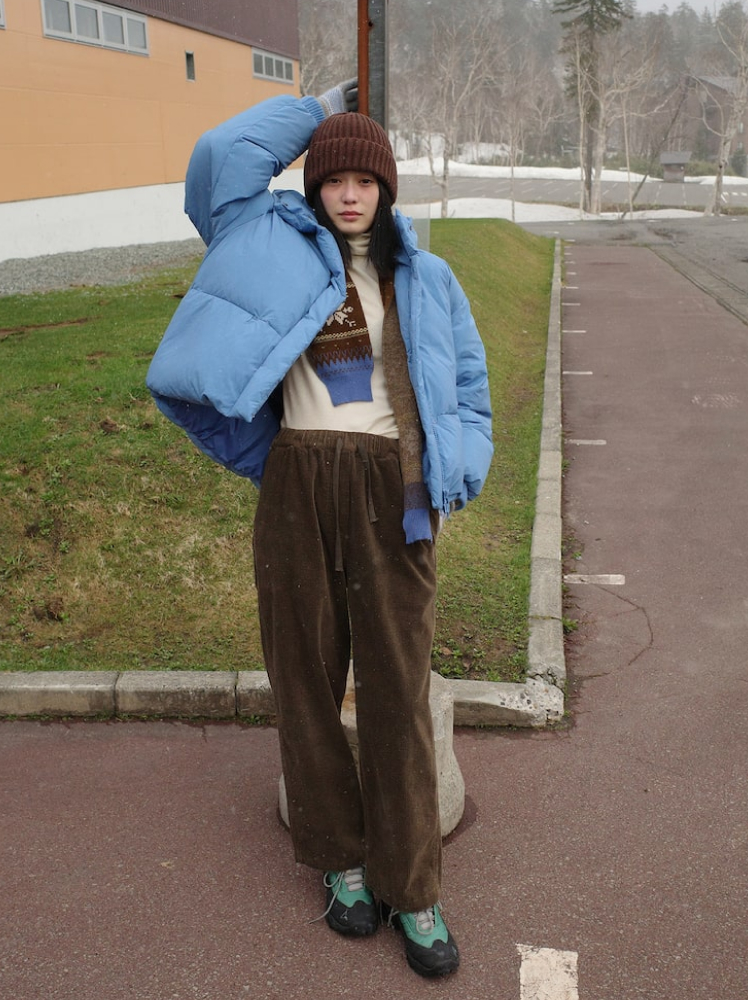 A woman poses with her hand on a street pole in a car park during winter and she wears a bright blue puffer jacket over a white turtleneck with brown corduroy trousers, hiking shoes, and a brown beanie and scarf.