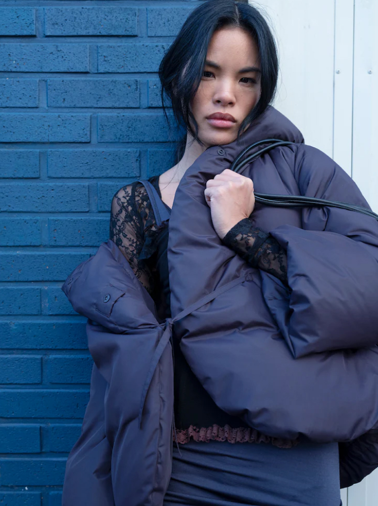 A woman leans up against a blue brick wall with a grey puffer jacket hanging off her shoulder, revealing a light knit mesh top underneath. She is holding her hand in a fist on her chest while clasping at the straps of her leather bag.