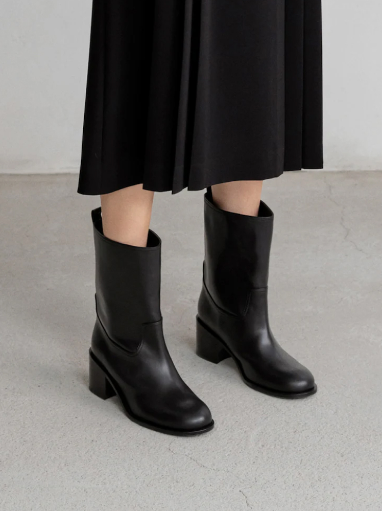 A model stands at a 45-degree angle to the camera wearing a pair of mid-calf black wide ankle boots with a small block heel; the boots are paired with a flowy black midi skirt.