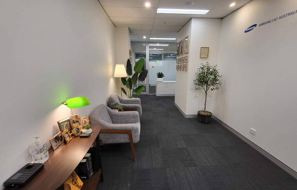 An image of the entry to the Samsung C&T Sydney branch office with grey carpeted flooring, grey arm chairs, several indoor plants, and a side table at the front door with photo frames with images of team member's pets from the office.