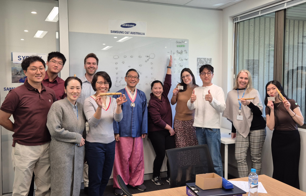 Eleven team members stand together in an office meeting room to take a photo after playing a traditional Korean game to celebrate the harvest festival season.