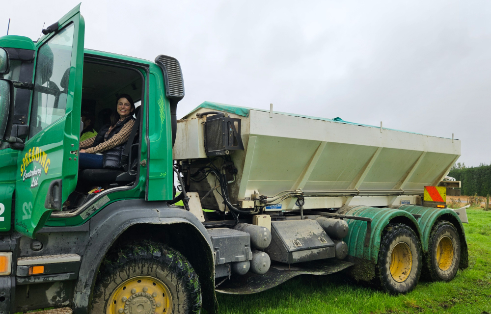 A woman smiles while sitting in the passenger's seat of a large green truck that is designed for spreading fertilizer on farmland.