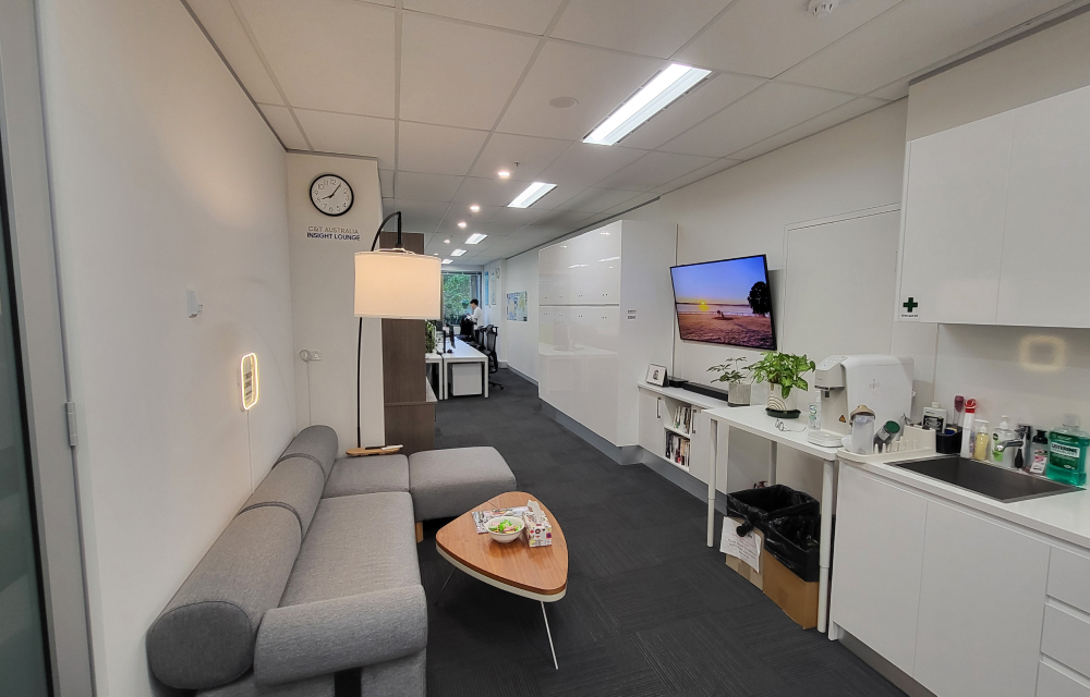 An image of the "Insight Lounge" at Samsung C&T's Sydney branch; the lounge consists of a grey sofa with a coffee table, a TV that hangs on the wall, and a kitchen with coffee available.