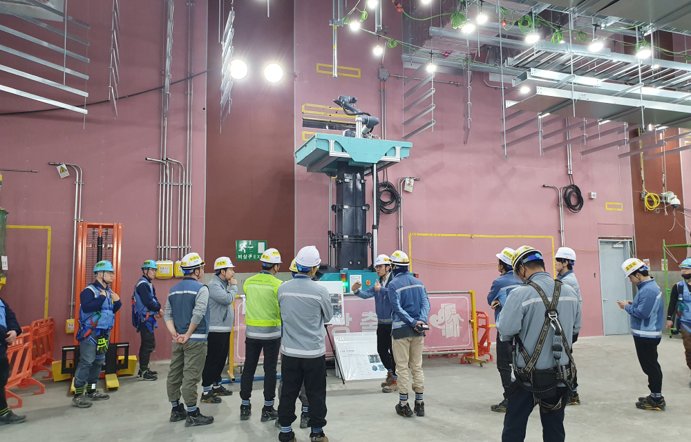 Construction workers wearing safety helmets and vests observe the Wall-Perforating Robot operating on-site. The environment includes structural components and equipment, showcasing advanced construction technology in a well-organized setting..