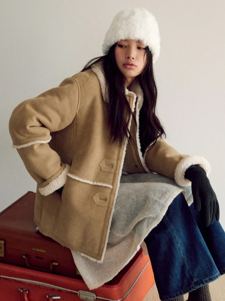 A model is seated on a pile of vintage suitcases while wearing a shearling jacket, jeans, gloves and a fuzzy hat for fall.