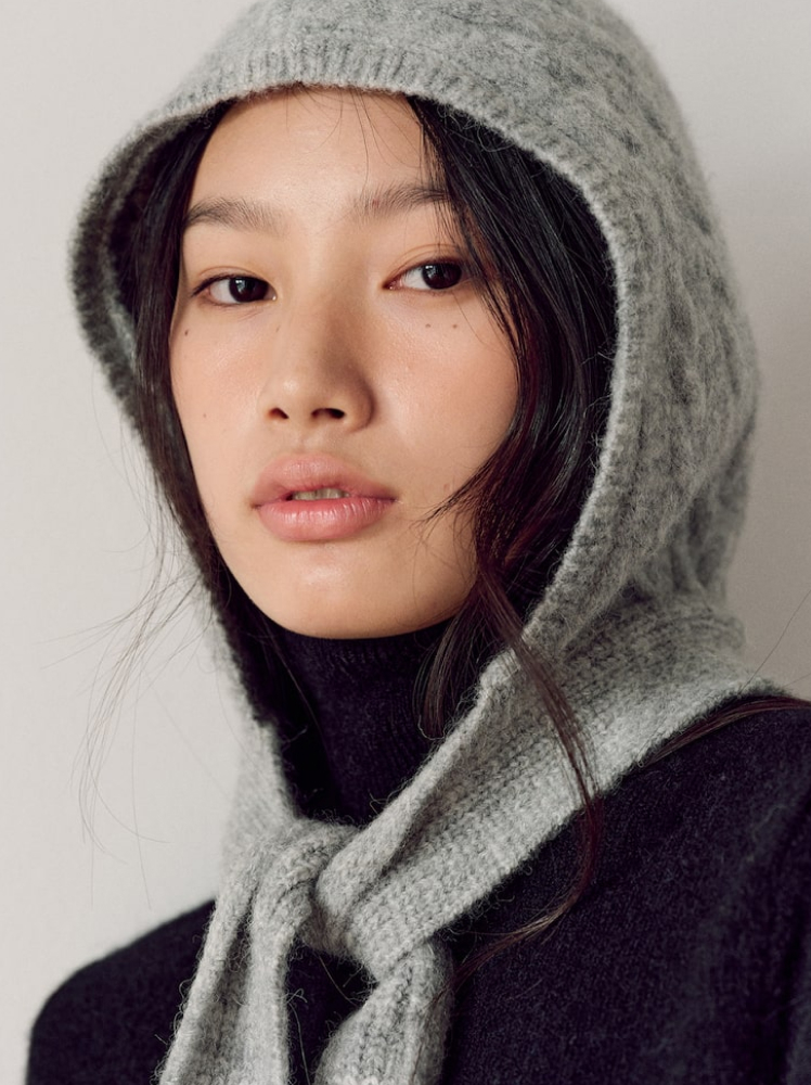 A model looks into the camera with a grey cable knit balaclava on her head with the thick straps of the hat tied around her neck like a scarf, over the top of her black turtleneck sweater.