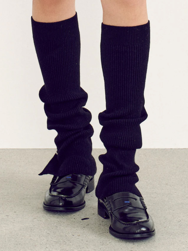 An image of a model’s legs wearing a pair of black knee-length leg warmers over a pair of patent leather loafers.