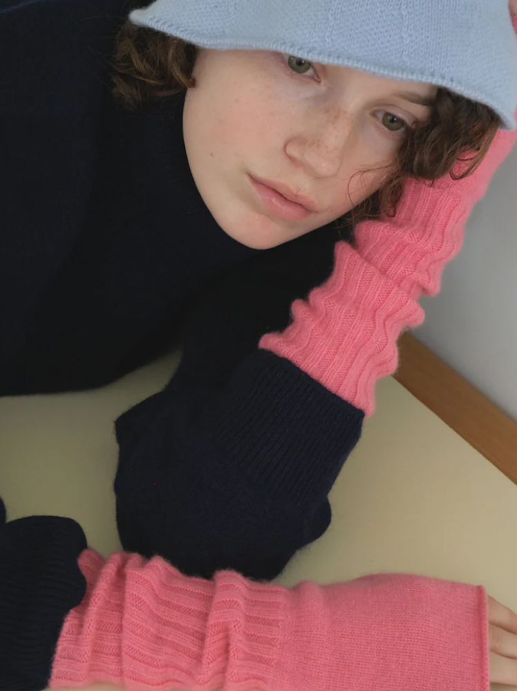 A model lays on the floor resting her head on her hands which have long length hot pink cut-off wool gloves; the model is wearing a pale blue knit bucket hat and a thick black turtleneck sweater.