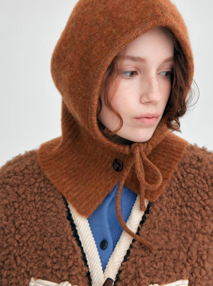 A model looks to her left while wearing a knit balaclava in a mustard brown color with button and tie up details at the neck. The balaclava is paired with a brown shearling jacket with a white trim on the collar and a cerulean cardigan underneath.