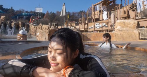 A visitor at Caribbean Bay rests against the edge of an outdoor spa pool, in the background is a large-scale outdoor adventure playground that has been transformed into an outdoor winter spa.