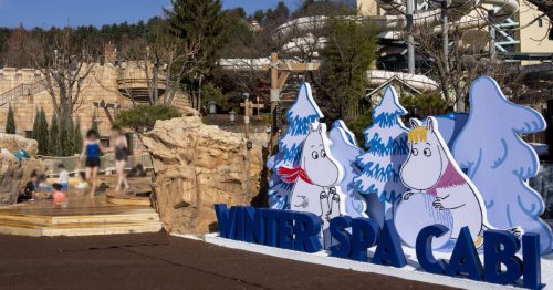 Card cut-outs of Moomin and Snorkmaiden rolling up a giant snowball sit behind a blue letter sign that read “Winter Spa Cabi” in front of outdoor spa pools and a giant outdoor water slide in the background.