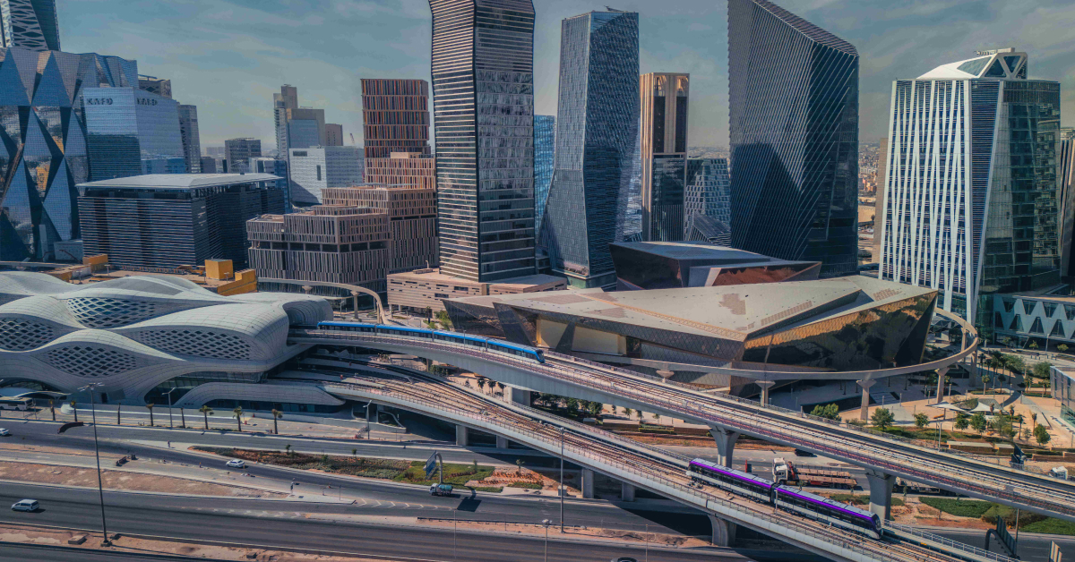 A vibrant urban skyline in Riyadh featuring modern architectural designs with high-rise buildings and a futuristic metro station. A metro train is visible on elevated tracks.