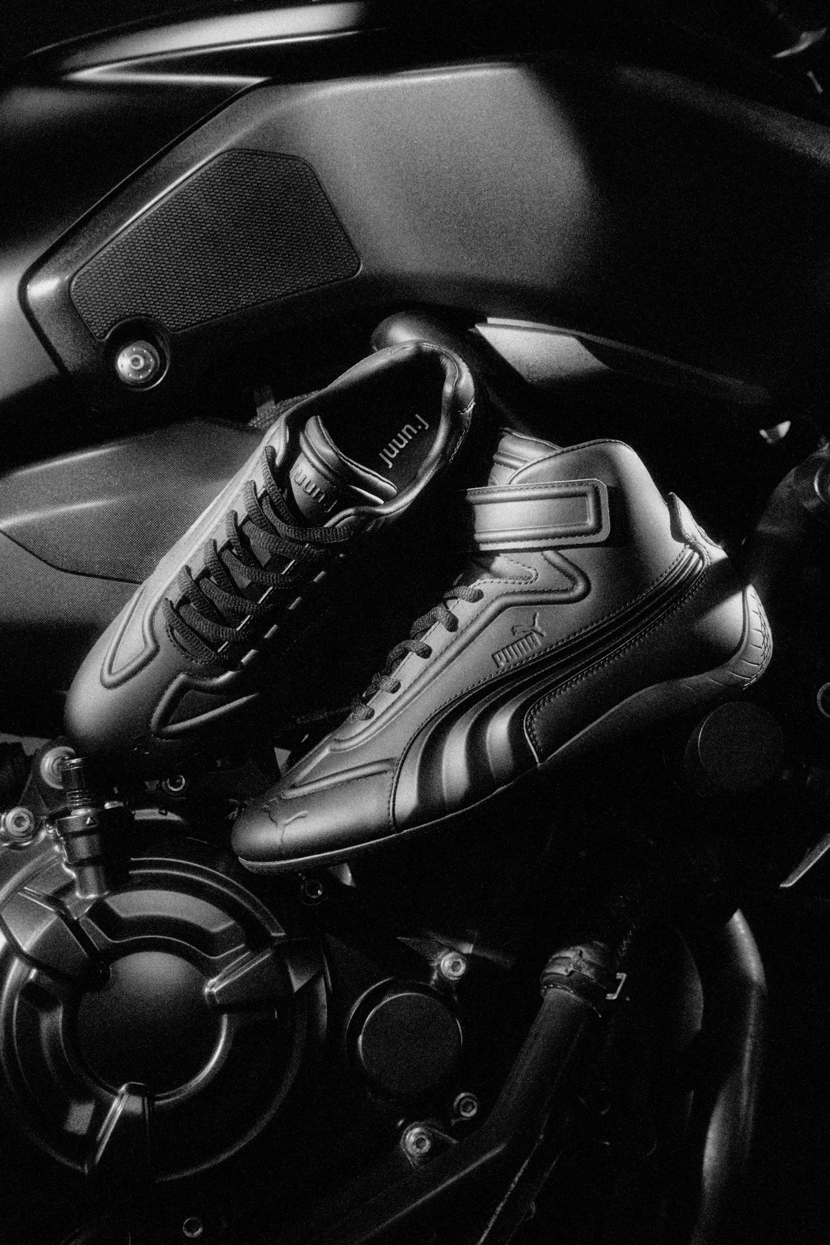 A close-up of PUMA Speedcat shoes in matte black leather, designed in collaboration with Juun.J, placed on a motorcycle engine, highlighting their minimalist and bold aesthetic.