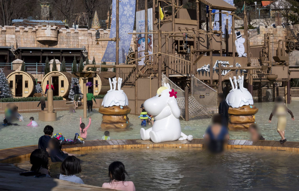 A wide shot of the outdoor adventure spa at Caribbean Bay’s winter spa; in the foreground, guests are seated in an outdoor spa pool next to Moomin characters including Snorkmaiden and the Hattifatteners, in the background is an aventure playground and three outdoor wooden barrel saunas.