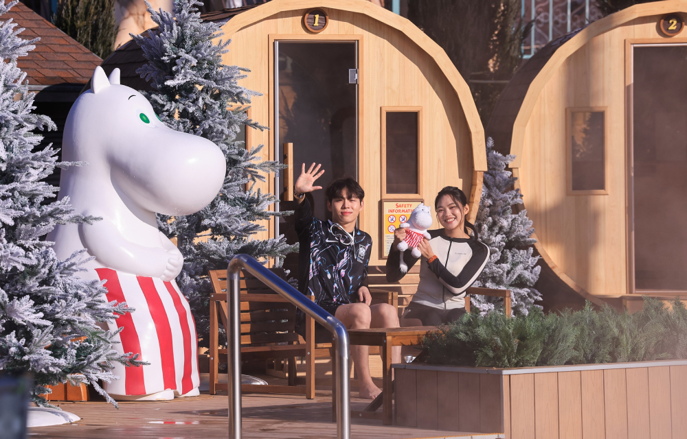 Two spa guests pose with a sculpture of Moominmamma on a wooden bench and are seated in front of a row of large wooden barrel saunas at an outdoor winter water spa and snow-covered Christmas trees.