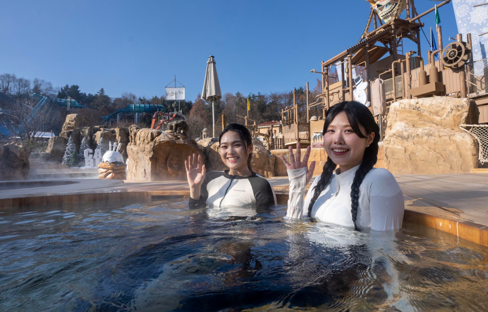 Two visitors wave at the camera while seated in an outdoor spa pool, in the background is the adventure pool at Caribbean Bay which has been transformed into an outdoor spa for the winter.