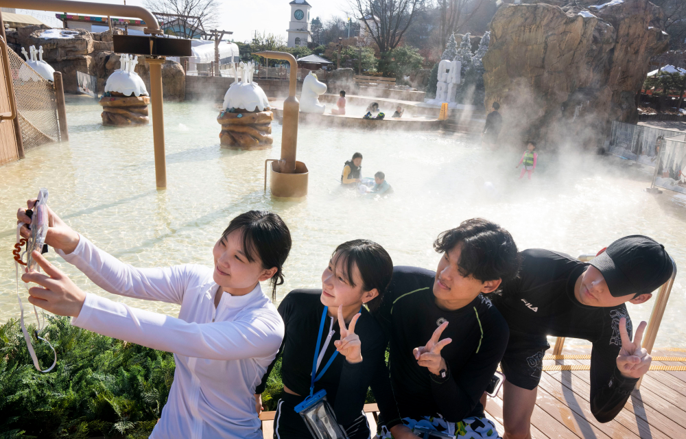 A group of 4 friends pose for a selfie while doing peace signs. In the background is a winter spa pool with steam from the warm water visible. In the very background are characters from the Moomin books.