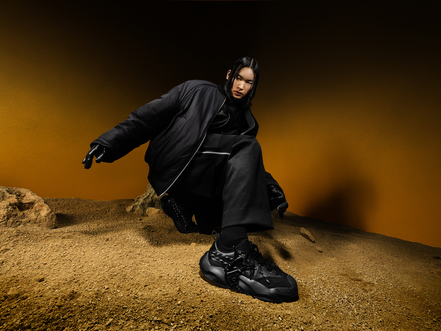 A model wearing the Juun.J x PUMA PLEXUS shoes poses in a Mars-like desert setting with an orange gradient background. The outfit features an oversized black jacket and wide pants, emphasizing sleek, futuristic styling. The shoes are chunky, black, and rugged, designed for both performance and fashion.