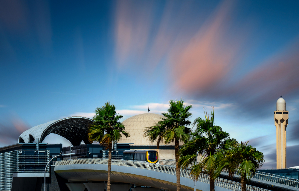 A view of a Riyadh Metro station near a domed structure, surrounded by palm trees. The scene highlights the blend of modern transit systems with the city's cultural heritage under a colorful, serene sky.