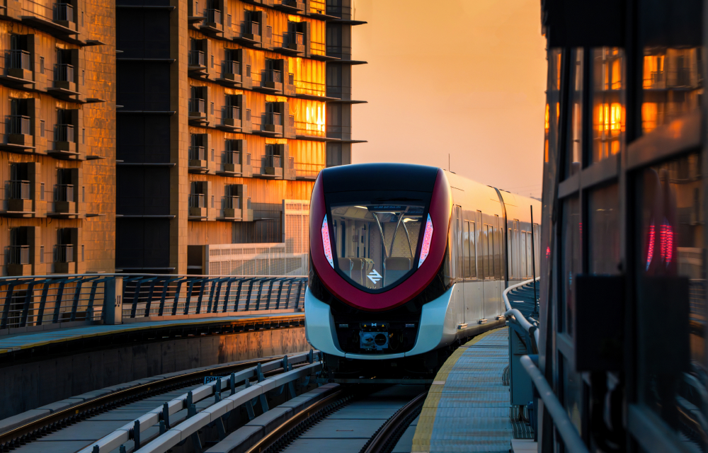 A new train along the Riyadh Metro system in Saudi Arabia emerges at sunset between 2 high rise buildings with the sunset glimmering in the windows.