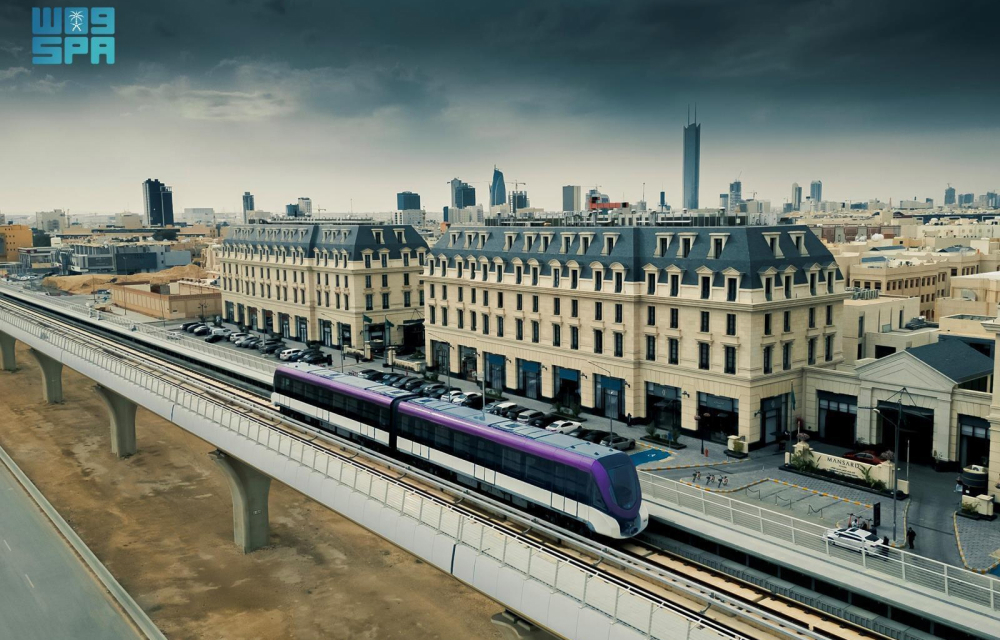 An elevated metro track with a modern train running alongside European-style buildings. The backdrop includes an urban cityscape with skyscrapers under a cloudy sky.