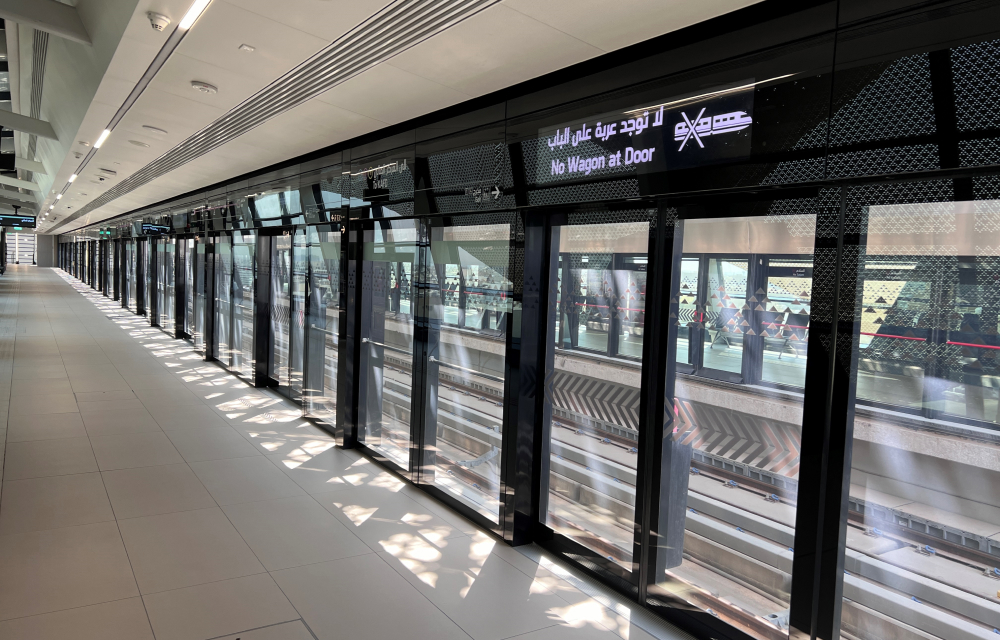 The interior of a Riyadh Metro station featuring a sleek, modern design with polished reflective walls and digital signage displaying train schedules and directions to various destinations.