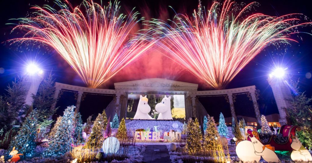 A nighttime view of Everland's Moomin Multimedia Fireworks Show featuring colorful pyrotechnics over a Nordic Forest setting with Moomin characters displayed on a large screen and festive lights decorating the area.