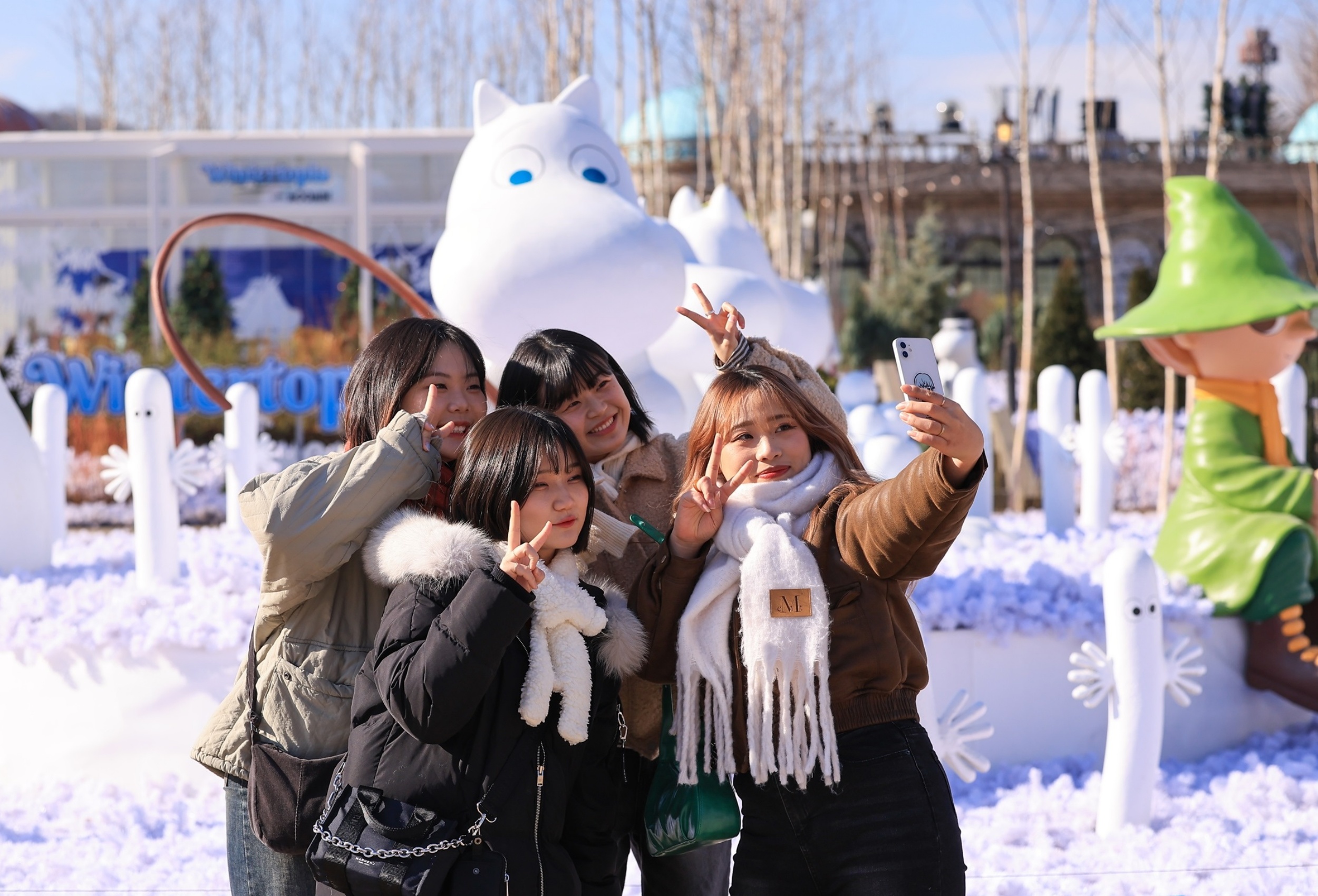 A group of friends pose for a selfie in front of the Moomin-themed Nordic Forest at Everland, surrounded by snowy decorations and Moomin characters under a bright winter sun.