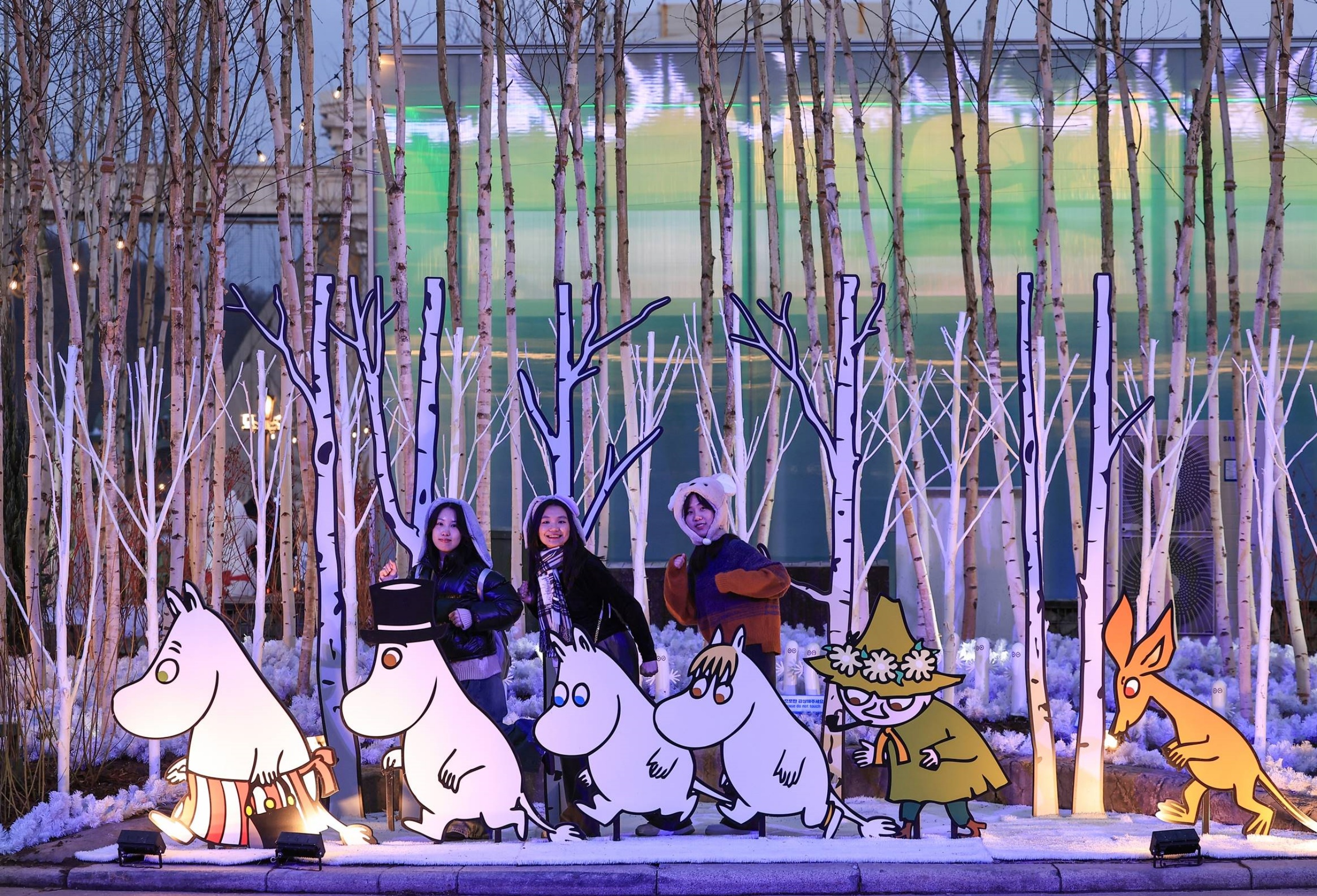 Visitors pose with illuminated Moomin character cutouts amidst a birch tree-themed display at Everland's Nordic Forest during nighttime.