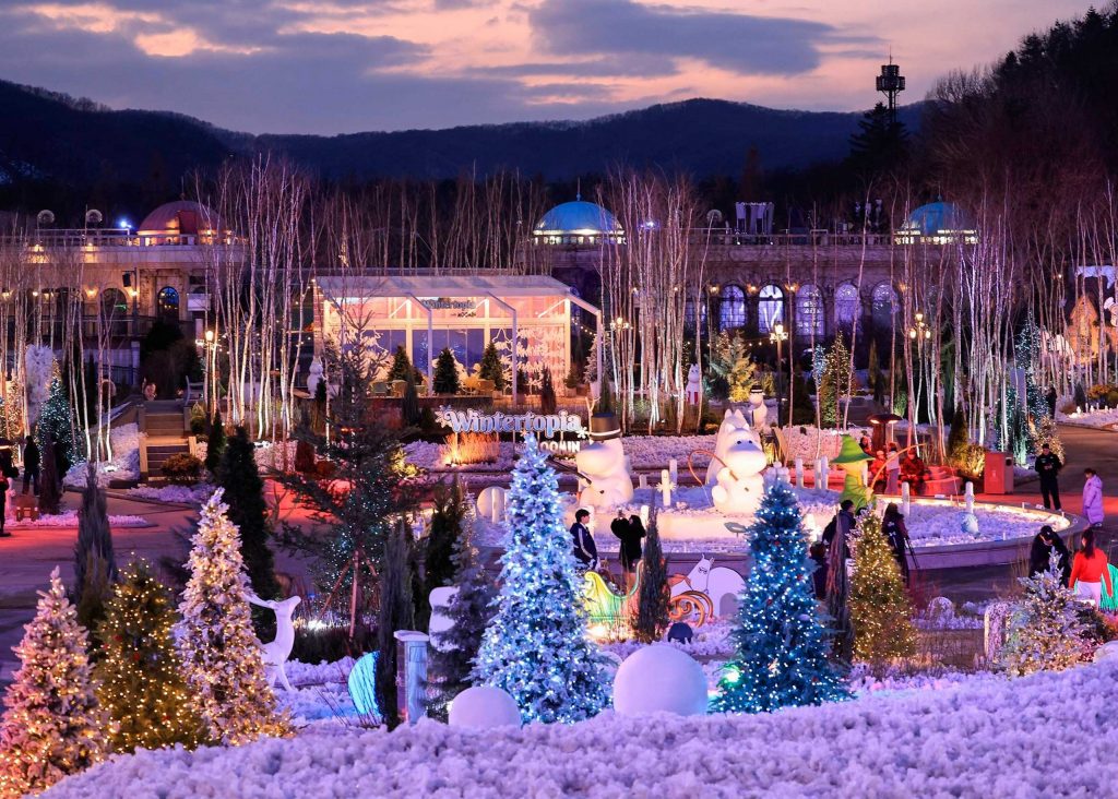 A picturesque view of Everland's Wintertopia during dusk, showcasing illuminated Christmas trees, Nordic-inspired decorations, and Moomin-themed sculptures amidst a snowy landscape. Visitors explore the festive area surrounded by glowing lights and a serene mountain backdrop.