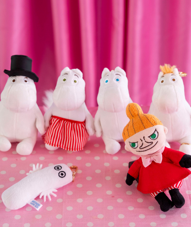 A collection of Moomin-themed merchandise displayed on a pink polka-dot table, featuring plush toys of Moomin characters and a white keychain with googly eyes.