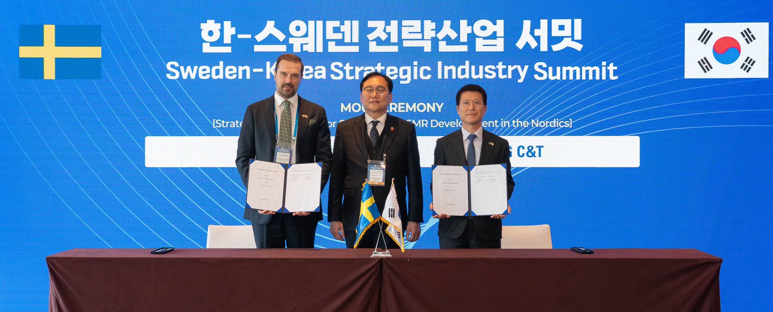 Samsung C&T and Kärnfull Next AB representatives holding signed documents at the Sweden-Korea Strategic Industry Summit, with flags of Sweden and Korea displayed on the table and event signage in the background.