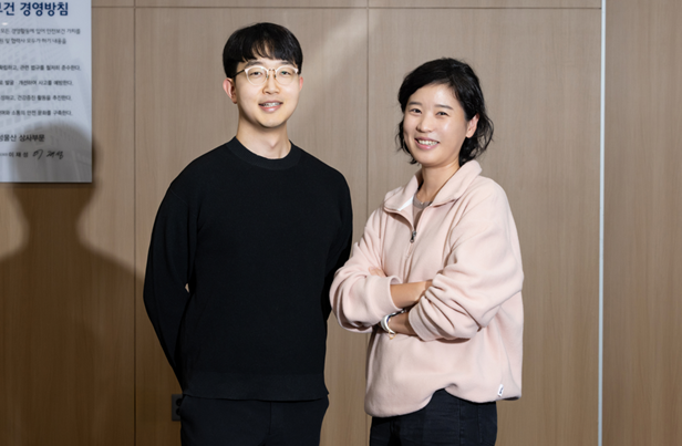 Two team members of the Digital Innovation Group, Seokyoon Jung in a black sweater and Yoonju Cho in a pink sweatshirt, standing side by side in an office setting.