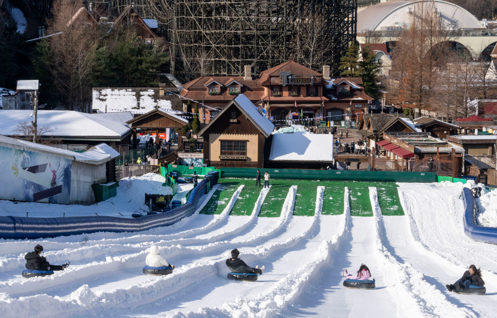 Visitors sledding down multiple snowy lanes of the Racing Course at Everland, with a backdrop of wooden roller coasters and charming cottages under a bright winter sky.