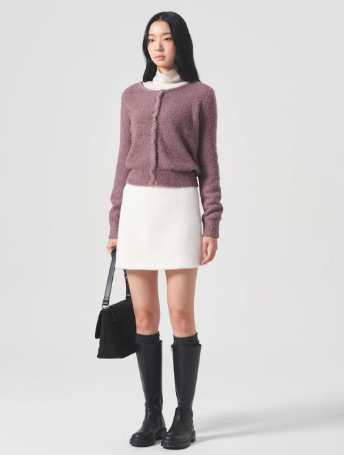 A woman in a fuzzy mauve cardigan, white mini skirt, and black knee-high boots, carrying a black bag.