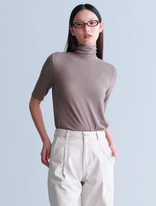 A woman wearing a taupe short-sleeve turtleneck tucked into high-waisted white trousers, accessorized with thin-framed glasses.