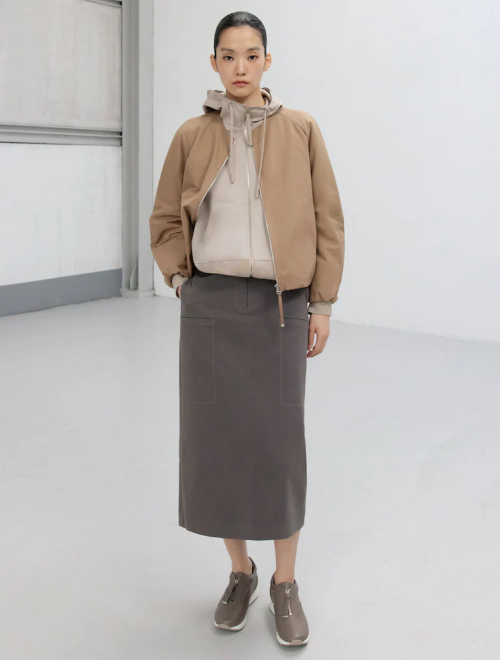 A woman in a mocha mousse bomber jacket over a taupe hoodie, paired with a long gray skirt and neutral sneakers.