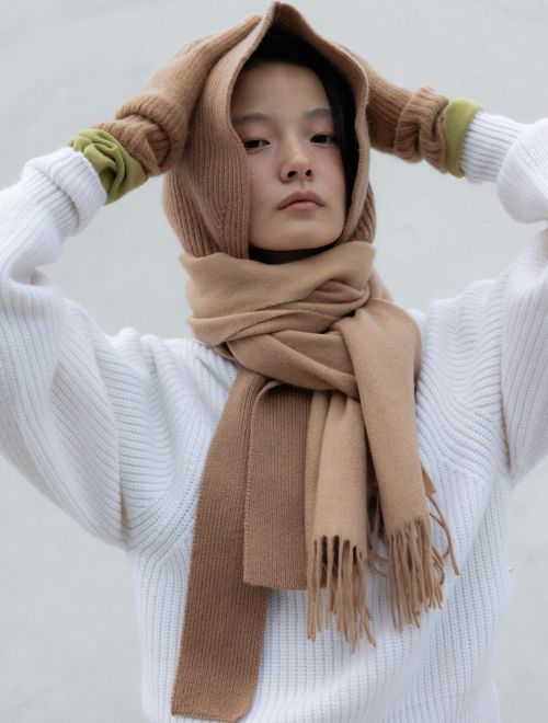 A woman wrapped in a camel-colored knit scarf and hood, layered over a white textured sweater with green gloves.