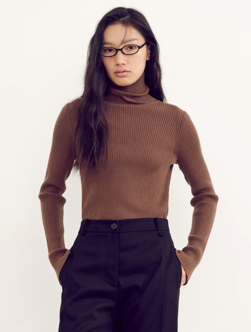 A woman in a ribbed brown turtleneck and black trousers, wearing small rectangular glasses.