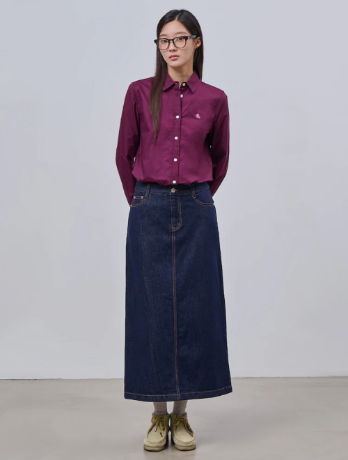 A woman in a button-up plum-colored shirt tucked into a long dark denim skirt, paired with glasses and beige shoes.