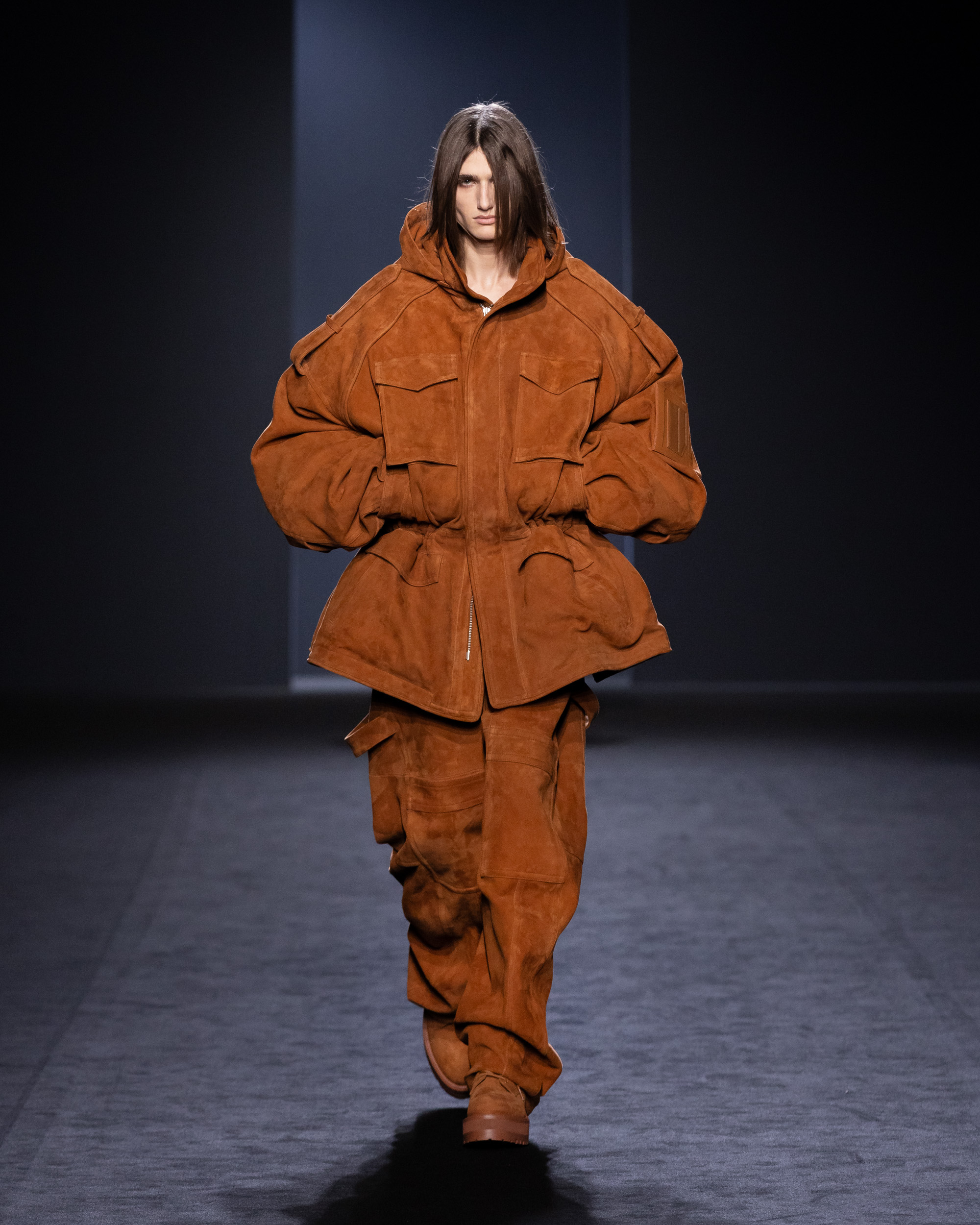 A model wearing an oversized rust-brown suede jacket with cargo-style details and matching pants walks the runway, reflecting Juun.J’s innovative blend of utility and luxury at Paris Fashion Week.