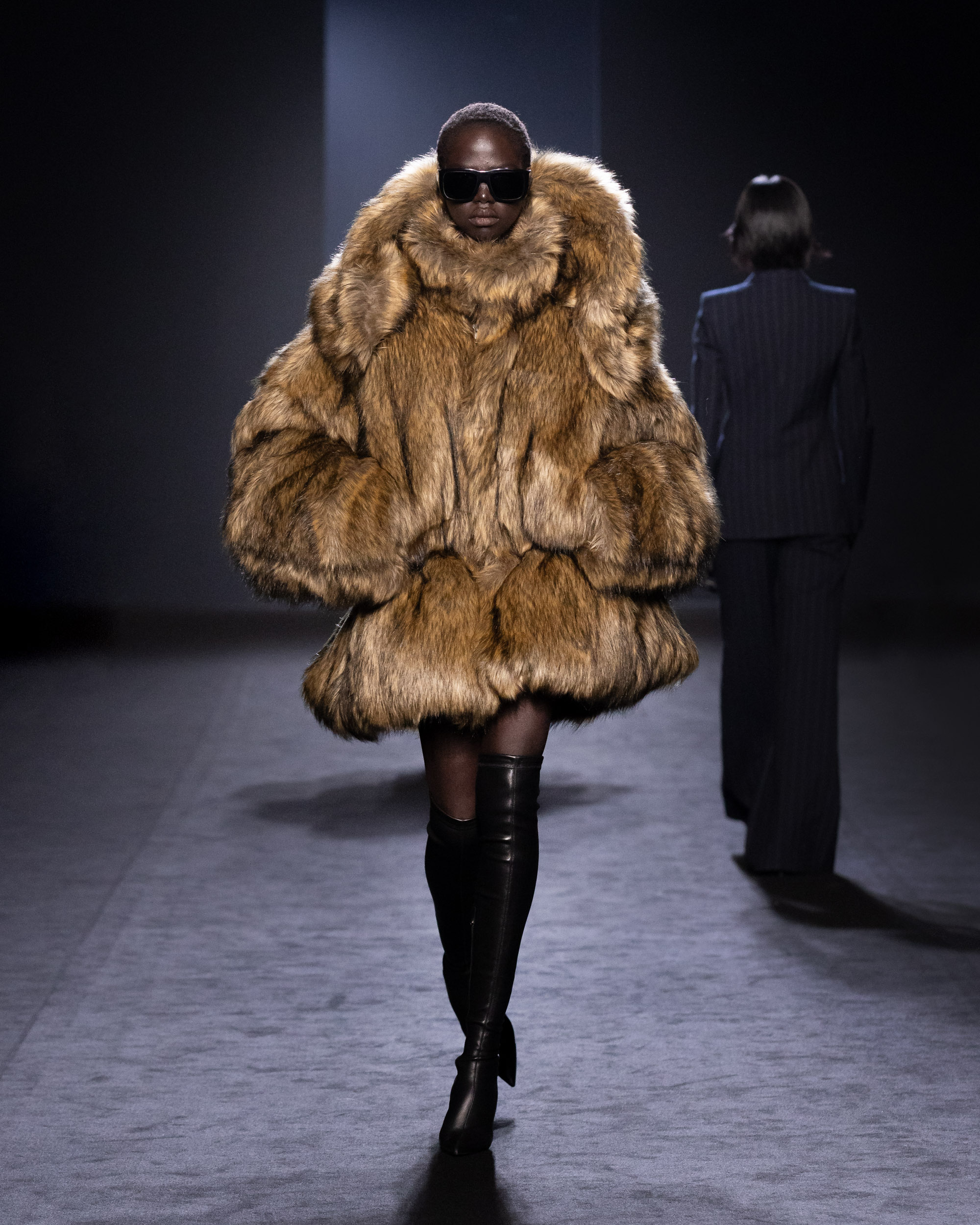 A model wearing a voluminous brown fur coat and thigh-high black leather boots walks the runway at Juun.J’s Fall/Winter 2025 Paris Fashion Week show, showcasing a bold and opulent look.