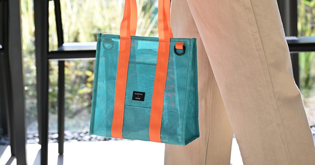 A turquoise tote bag with bright orange straps, part of Samsung C&T E&C Group’s upcycled accessories collection, made from construction waste. The bag is hanging from a person’s arm as they stand near a black bench outdoors. This design, which won an iF Design Award, reflects Samsung C&T’s commitment to sustainability and innovative reuse of construction materials.