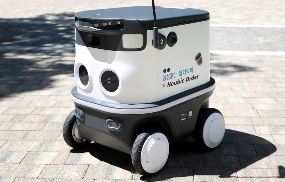 A four-wheeled autonomous delivery robot branded with "Samsung C&T Deli-Pick Me x Neubie Order" is positioned on a paved outdoor surface. The compact robot features sensors, cameras, and a sleek design optimized for safe navigation and efficient residential deliveries.