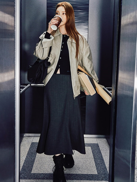 xooos is captured in an elevator wearing the "Minimal Leather Jacket Look" from the second drop of the 8seconds x xooos collaboration. She styles a beige Edition8 Oversized Leather Jacket with a navy ribbed cardigan, striped top skirt in ash, and a black nylon shoulder bag. Holding books and a coffee cup, her look reflects a contemporary city aesthetic with an understated yet polished charm.