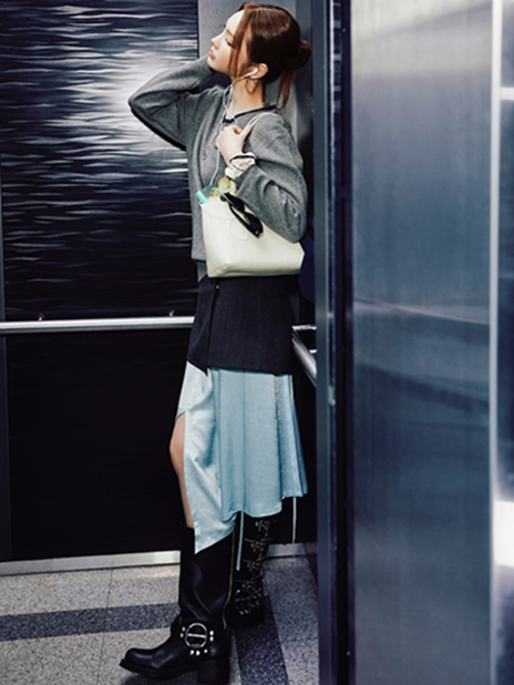 xooos poses in an elevator wearing the "Minimal Layered Look" from the second drop of the 8seconds x xooos collaboration. She is dressed in a grey Edition8 Double V-Neck Pullover, sky blue Edition8 Bias Cut Skirt, and accessorized with an ivory PU Shoulder Tote Bag while wearing black knee-high boots. The combination of knit textures and flowing layers creates a modern yet effortlessly feminine silhouette.