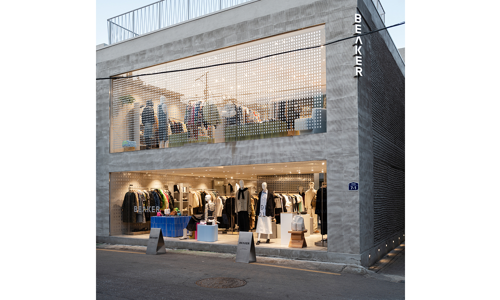 Exterior of BEAKER’s modern, industrial-style store featuring raw concrete walls and large glass windows. The two-story building showcases stylish clothing displays inside, with a clean and minimalist design that integrates seamlessly with the urban surroundings.