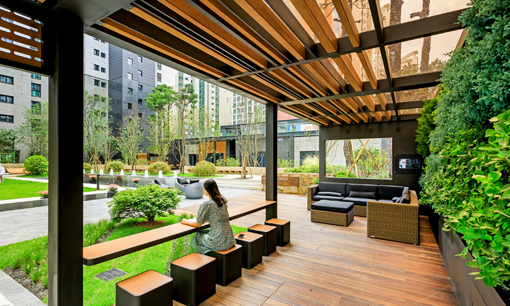 A shaded outdoor seating area under the Green Oasis Pergola at Raemian La Classe. The wooden pergola with slatted roofing provides a cozy retreat with cushioned lounge chairs and tables, surrounded by lush greenery and landscaped gardens, offering residents a relaxing escape in an eco-friendly environment.